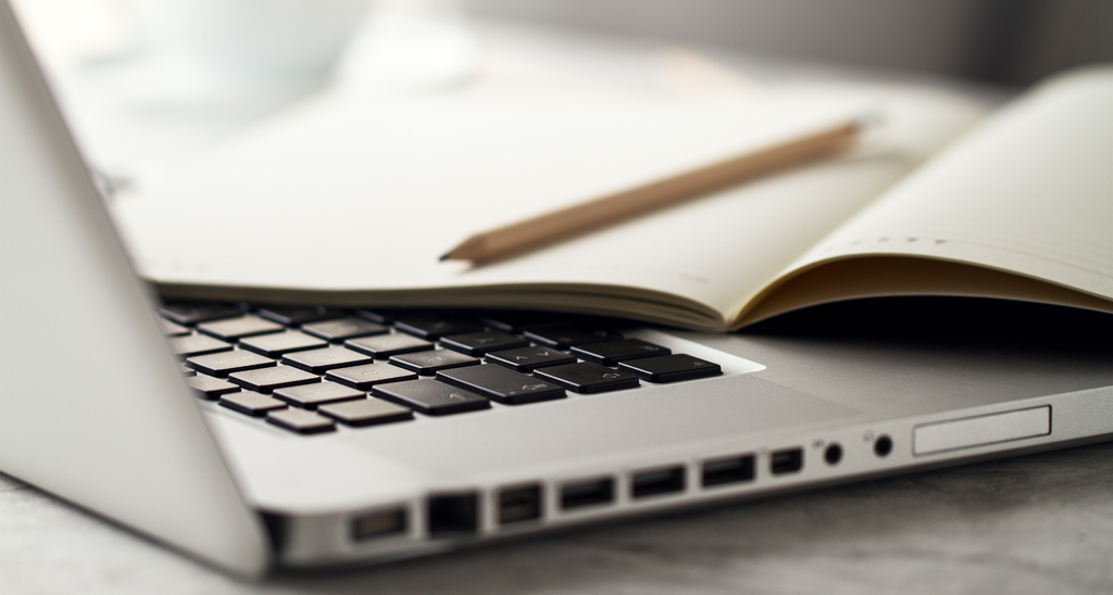 Foto de um computador, um lápis e um caderno.