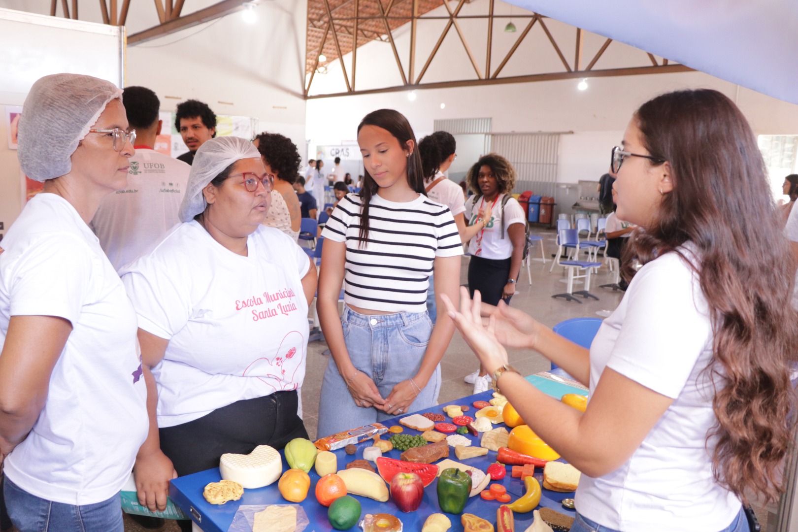 Imagem de várias pessoas estão reunidas em torno de uma mesa com alimentos diversos com estudante do curso de Nutrição da UFOB fazendo apresentação durante 1ª edição do Programa UFOB no Bairros, no dia 10 de maio de 2025, em Barreiras