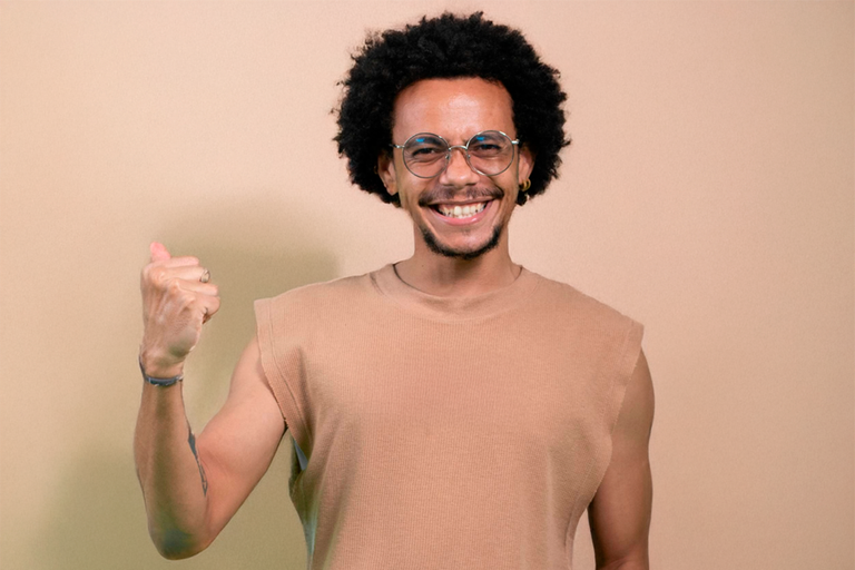 Jovem negro, com camiseta marrom, sem mangas, fazendo gesto que simboliza vitória e conquista, com os punhos cerrados.