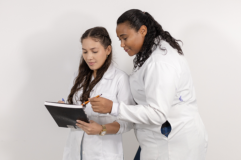 Imagem de duas estudantes, uma está com caderno na mão e a outra olhando o que está escrito nele