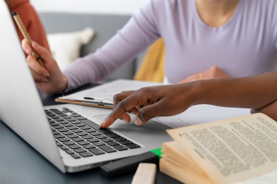 Imagem de duas pessoas em frente ao notebook, com a mão de uma no teclado e um livro ao lado
