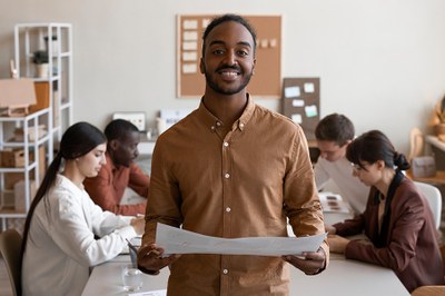 Imagem de professor negro em destaque com equipe sentada em mesa ao fundo