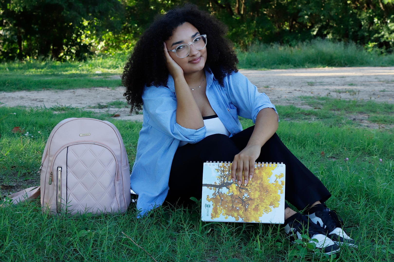 Imagem de estudante da UFOB sentada numa área verde com o caderno da Universidade em uma das mãos