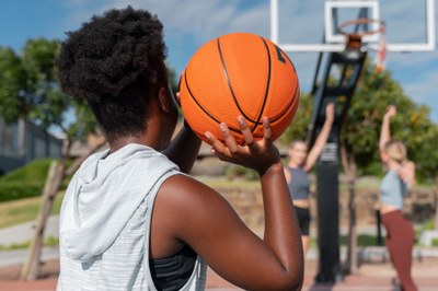 Imagem de três mulheres jogando basquete, a primeira com a bola na mão em posição de arremesso e as outras duas logo abaixo da tabela