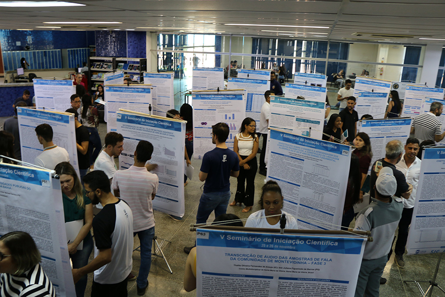 Imagem de estudantes em um ambiente fechado apresentando pôsteres com resultados de pesquisas de iniciação científica e tecnológica