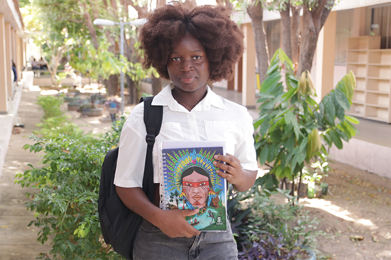 Foto de uma estudante do programa PEC-PG, de origem africana, segurando um caderno da UFOB, com um jardim ao fundo